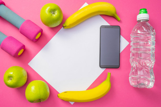 苹果、香蕉、一部手机、一瓶水和哑铃躺在普通的粉色背景上图片下载