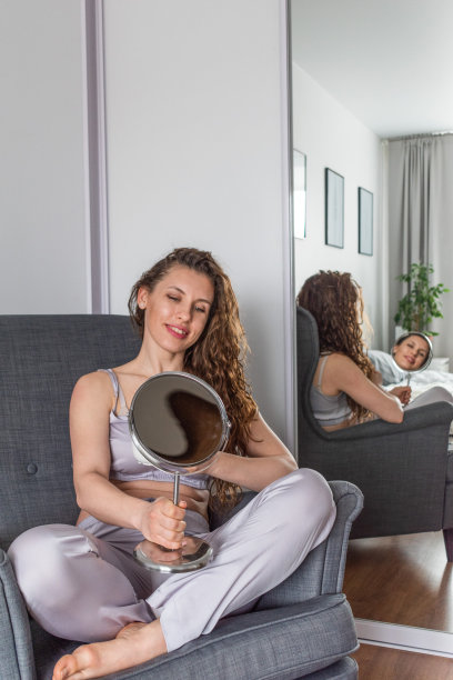 年轻女子坐在扶手椅上，拿着一面镜子看着自己。人类健康的概念。图片下载