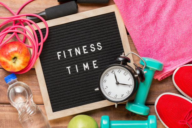 健身时间和运动概念。红色运动鞋，苹果，跳绳，哑铃，粉色毛巾，老式闹钟，木制背景上写着健身时间的黑色牌子。图片下载