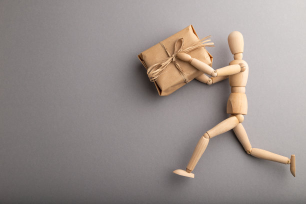 木制人体模型拿着一个礼盒在灰色柔和的背景。拷贝空间，隔离，交付概念。图片下载