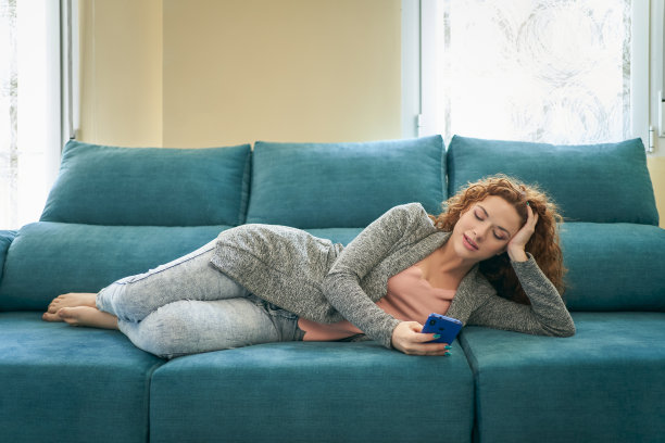 年轻的女博主躺在时尚公寓舒适的沙发上，通过智能手机在网上发布新帖子。时髦女孩通过电话在社交网络上阅读新闻图片下载