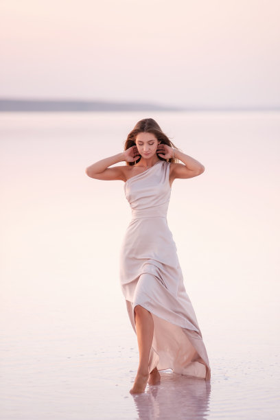 年轻的金发女郎穿着一件淡粉色的晚装，光着脚站在白色的结晶盐上。自然化了妆的女孩，头发正在生长。盐矿之旅，在夕阳下的水上行走图片下载