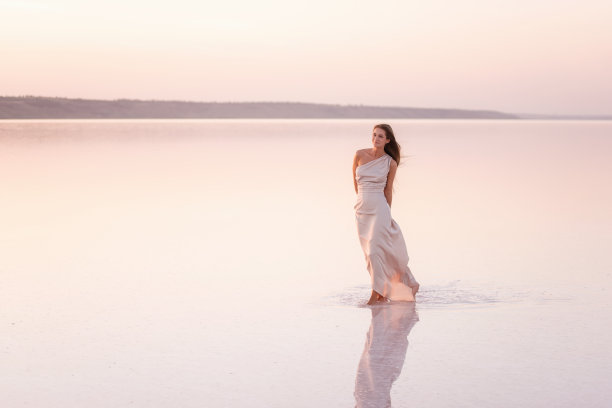 年轻的金发女郎穿着一件淡粉色的晚装，光着脚站在白色的结晶盐上。自然化了妆的女孩，头发正在生长。盐矿之旅，在夕阳下的水上行走图片下载