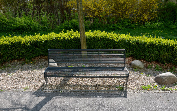 灰色金属公园长椅与透明金属线交叉杠的设计，在公园花坛的背景米色碎石路面休息区与黄杨图片下载