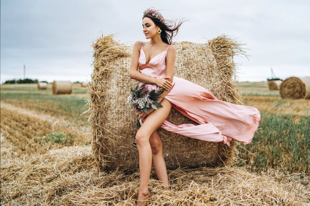 年轻的黑发穿着粉红色的缎子衣服，没有亚麻站在干草捆附近在大风天气。一个女人手里拿着一束野花图片下载