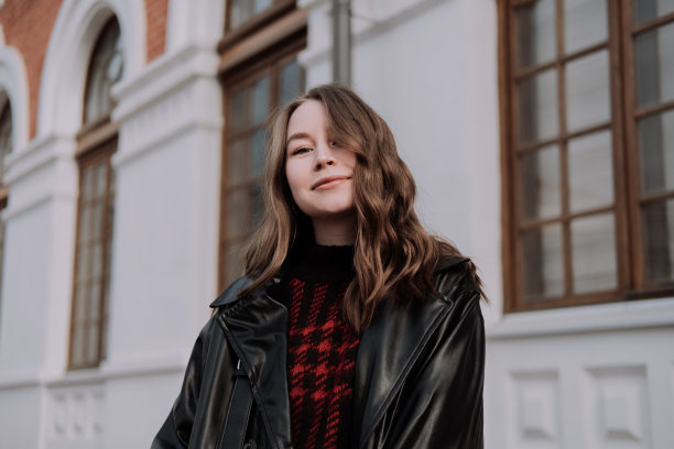微笑的年轻女子在黑色皮夹克和毛衣站在前面的白色墙壁看着相机。都市风格与街头时尚。穿着休闲装的卷发女孩图片下载