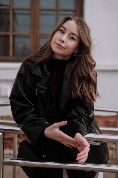 身穿黑色皮夹克的年轻女子微笑着站在白墙前，靠在铁栏杆上，看着摄像机。都市风格与街头时尚。穿着休闲装的卷发女孩图片下载