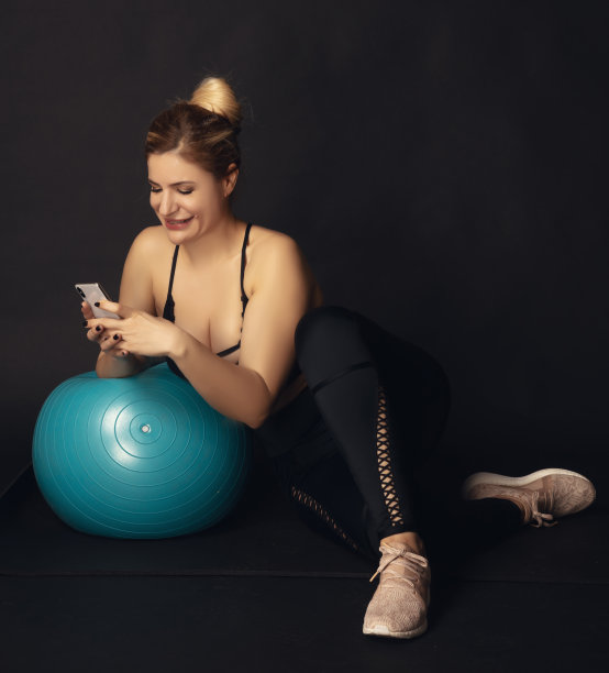 女运动员在锻炼时休息，坐在地板上靠在普拉提球上，同时在手机上发短信。图片下载