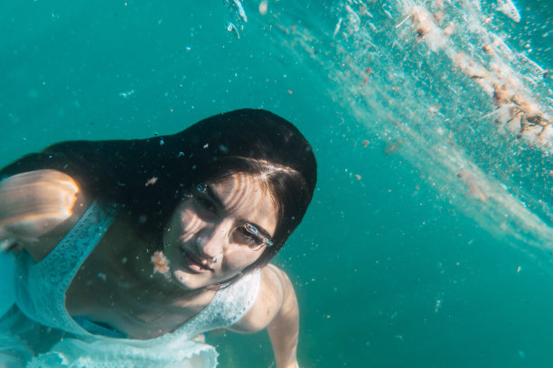 一个年轻的女人将潜水与白色的裙子游泳，水下的图像，夏天的生活方式，一个年轻的白人黑发看着相机图片下载