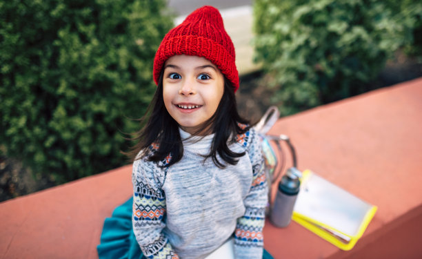 戴着红帽子的漂亮小女孩坐在学校旁边的户外。放学后，漂亮的孩子在外面休息。图片下载