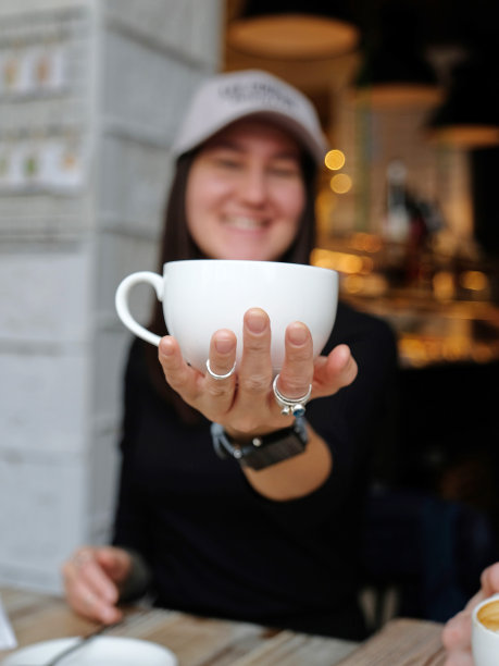 一个女孩坐在咖啡店里，手里拿着一个白色的咖啡杯。微笑。这张脸在背景中，焦点模糊。图片下载