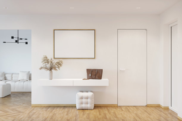 白色的入口走廊上挂着一幅空白的水平海报，上面是一个柜子，花瓶里插着潘帕斯草，镶木地板上放着一个蒲团，有两扇门，一扇门通向配有白色沙发的客厅。图片下载