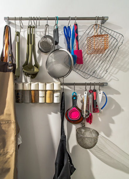 挂着厨房工具、厨房用具、围裙和调味品的金属栏杆在厨房的白色墙壁前。图片下载