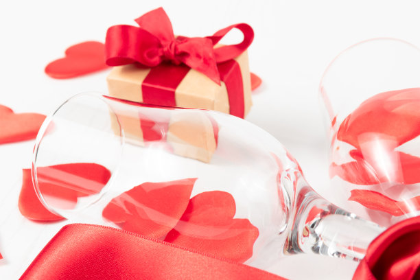 白色木桌上摆放着两个香槟杯，一个礼盒，一条红色的绸带和许多爱心图片下载