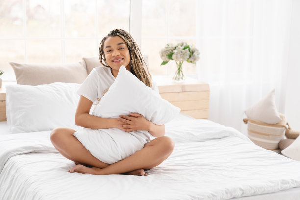 快活的年轻非洲妇女抱着一个枕头，坐在床上，在起床前放松。白色床单，亚麻布和整形床垫图片下载