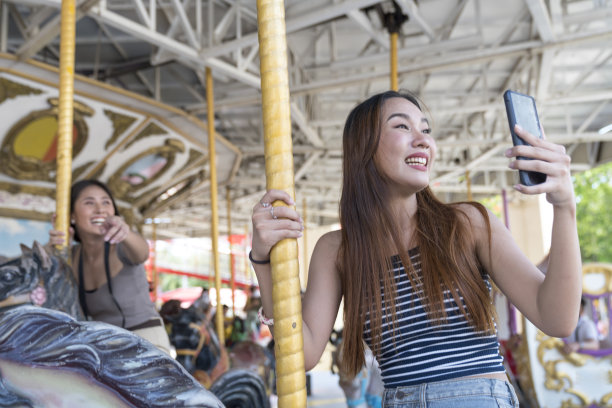 年轻漂亮的女人在游乐场的旋转木马上自拍。两个迷人的女人在游乐场自拍。图片下载