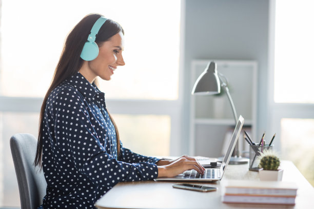 迷人的年轻女子的侧面照片写笔记本电脑坐办公桌戴耳机办公室工作人员在室内工作图片下载