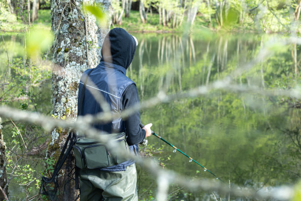 年轻的渔民在他的背部在运动衫和帽衫与背心和篮子与钓具持有杆旁边的水和拷贝空间。图片下载