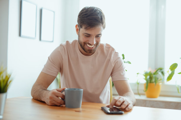 英俊的男人与完美的牙齿微笑和使用智能手机，而喝咖啡图片下载