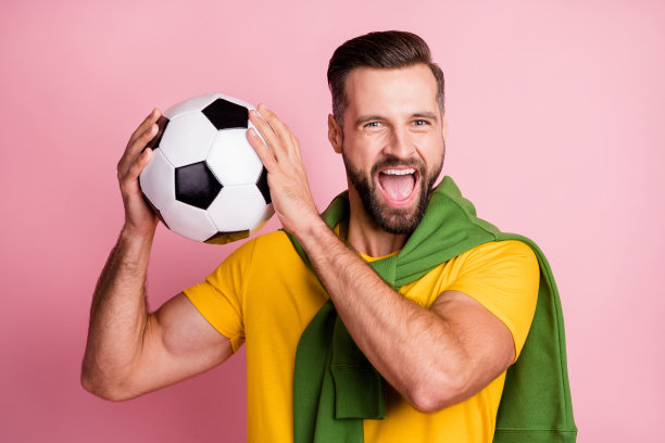 照片的棕发英俊的男人抱球穿套头衫在肩膀上足球球迷孤立在柔和的粉红色背景图片下载