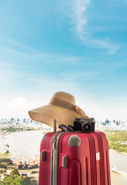 度假和旅行。带帽子的旅行箱或行李袋，相机和太阳眼镜，以城市和河景为背景。图片下载