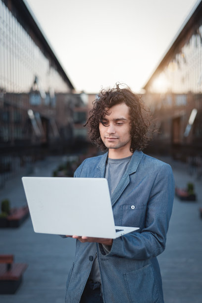 一个穿着夹克的英俊的年轻人，在一个现代化的商业中心或校园里，在一台白色的笔记本电脑前工作。学生、IT创业企业家或自由职业者。垂直的照片。图片下载