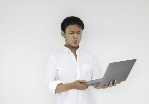 哇脸的年轻亚洲人震惊了他看到的笔记本电脑时，穿着白衬衫工作孤立的灰色背景图片下载