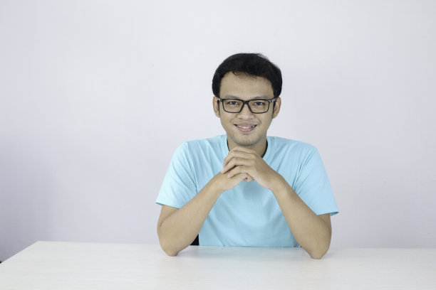 年轻的亚洲男子穿着蓝色衬衫，戴着眼镜，脸上挂着幸福的笑容，手托下巴放在桌子上。图片下载