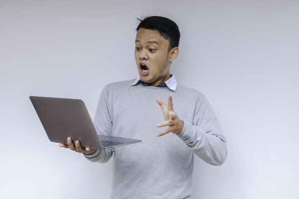 哇脸的年轻亚洲人震惊了他看到的笔记本电脑时，穿着灰色衬衫工作孤立的灰色背景。图片下载