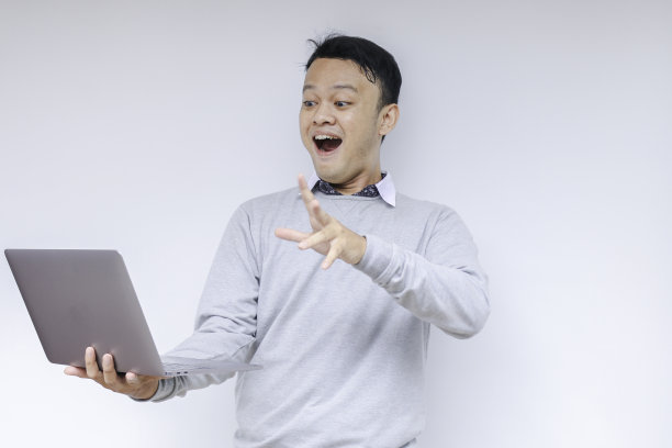 哇脸的年轻亚洲人震惊了他看到的笔记本电脑时，穿着灰色衬衫工作孤立的灰色背景。图片下载