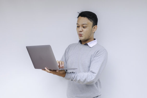 哇脸的年轻亚洲人震惊了他看到的笔记本电脑时，穿着灰色衬衫工作孤立的灰色背景。图片下载