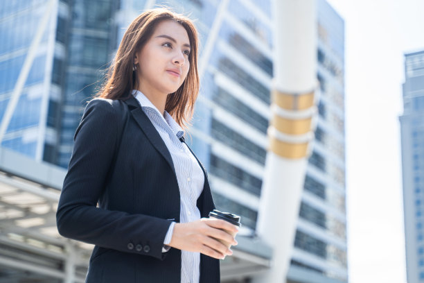 亚洲年轻的职场女性站在城市的户外，面带自信，端着一杯热咖啡享受生活。女工喝完茶点饮料后，心情舒畅，微笑着。图片下载