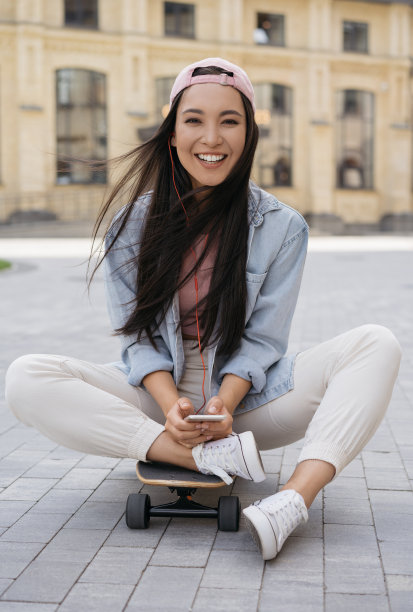微笑的亚洲妇女使用手机，听音乐在街上。快乐的情绪青少年坐在滑板上看相机图片下载