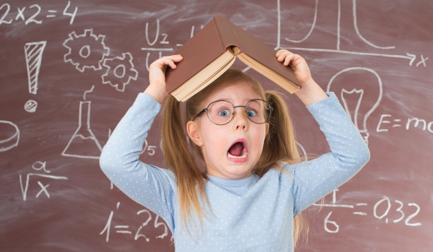 这是一个漂亮的女学生戴眼镜的特写照片，她把一本书举过头顶，因为学习和玩累了。戴眼镜、尖叫的小女孩，旁边是写满符号和公式的黑板。教育的概念。图片下载