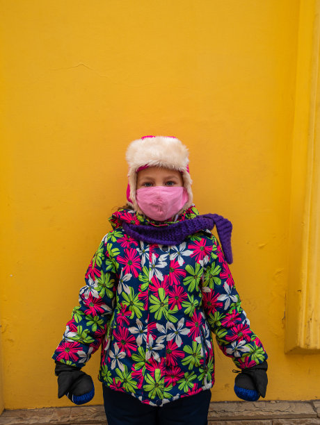 年轻的女孩在温暖的冬天的衣服与保护粉色面膜在黄色的墙壁背景。城市风格街景照片。图片下载