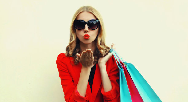 美丽的年轻女子的肖像与购物袋吹她的红色嘴唇送吻穿着红色的商务西装在背景图片下载