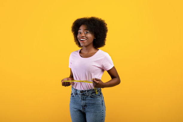 减肥的概念。适合非洲裔美国女士测量她的腰与胶带在黄色演播室背景图片下载