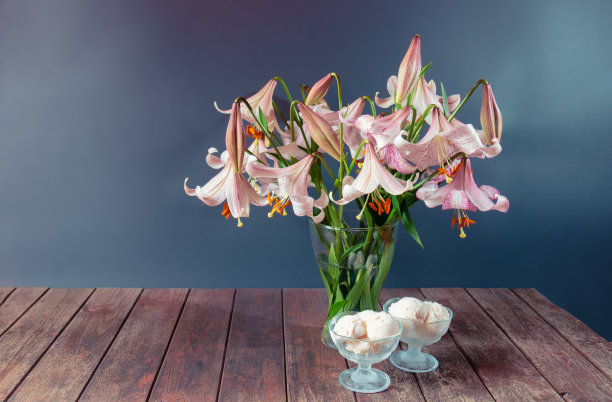 灰色背景的木桌上放着一束浅褐色带粉红斑点的百合花和几碗冰淇淋。图片下载