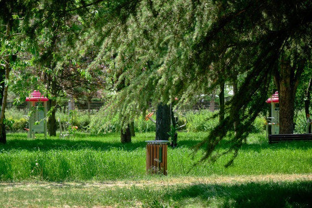 在阳光明媚的日子里，在布尔萨的一个绿色公共和自然公园里的废纸篓和垃圾桶，背景是操场。绿色草地上覆盖着木质材料的垃圾桶。图片下载