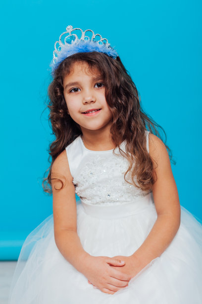 美丽的小卷曲的女孩与皇冠皇冠在白色连衣裙上的蓝色背景图片下载