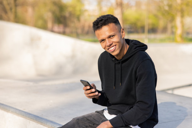 一个皮肤黝黑的男孩在公园里等着，手里拿着手机写着一条信息，他微笑着看了看向他走来的女孩，眼睛里闪烁着光芒图片下载