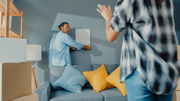 快乐的亚洲年轻迷人的男女互相帮助在墙上挂画框装饰房子和纸箱包装在客厅。图片下载