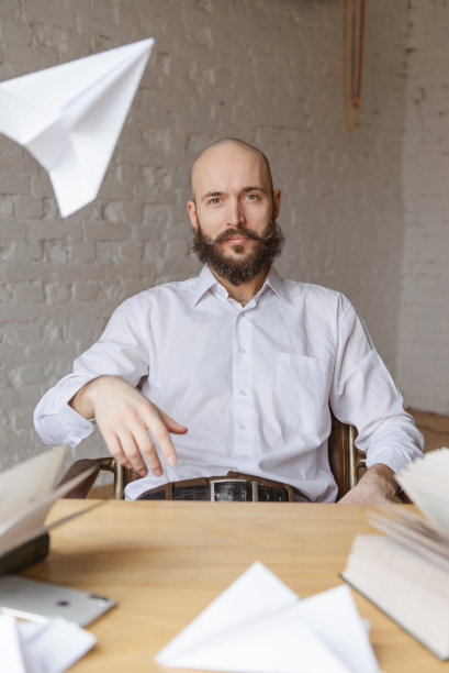 一个留着胡子、穿着白衬衫的白头男人正坐在一张桌子前飞纸飞机图片下载