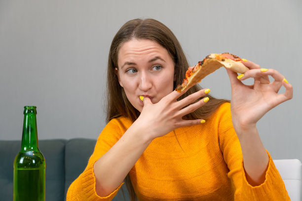 一位穿着橙色运动衫的漂亮的黑发年轻女子一手拿着一块披萨，另一只手的手指在舔着，旁边是一瓶绿色瓶子里的啤酒。快餐的概念图片下载