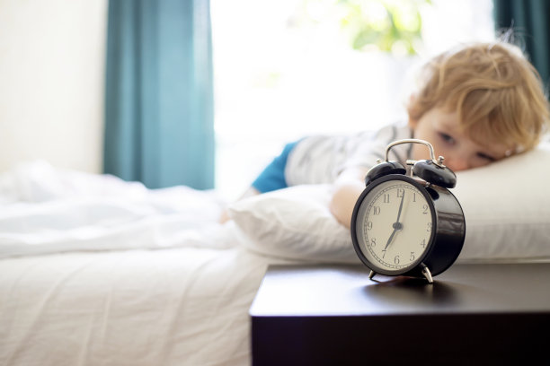 瞌睡的小孩躺在床上，伸手去拿响了的闹钟。瞌睡的男孩在床上，闹钟在前面。图片下载