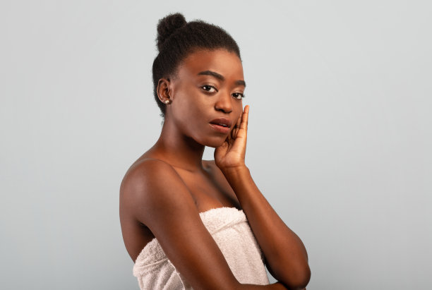非洲的美丽。漂亮的黑人女士裹在毛巾洗澡后，触摸她的下巴，在灰色的背景相机摆姿势图片下载