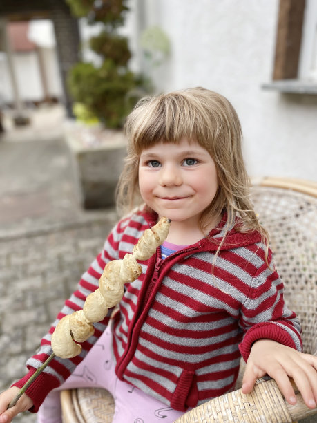 一个蹒跚学步的小女孩拿着一串面包，在火上烤着。有趣的快乐的孩子咬和吃受欢迎的聚会和营地烧烤食物在德国称为斯托克布罗特。图片下载