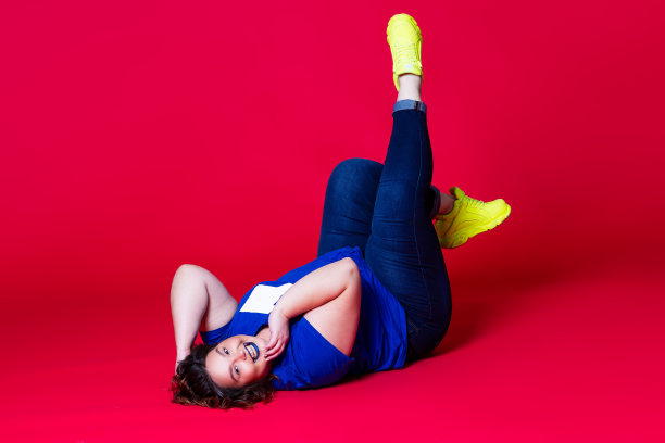 快乐加码模特在休闲衣服，胖女人在蓝色牛仔裤，t恤和黄色运动鞋在红色的背景图片下载