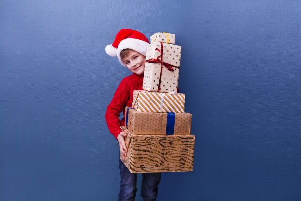 小男孩戴着圣诞帽，蓝色背景是圣诞礼物盒。圣诞购物。副本的空间。图片下载