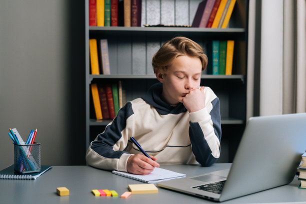 一个疲惫的小学生用钢笔在笔记本上做笔记，坐在书桌上用笔记本电脑，闭上眼睛。图片下载